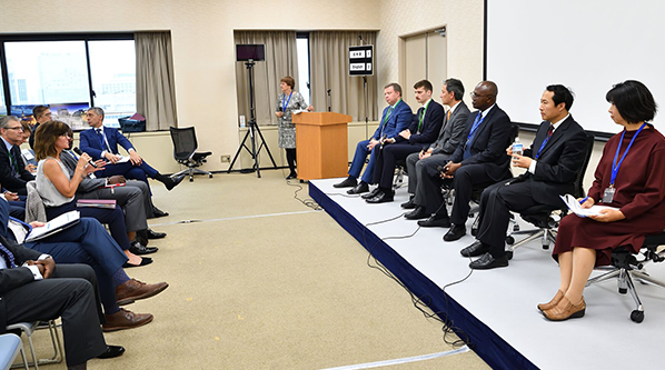 woman at podium and six events panelists seated in front of the front row of audience members